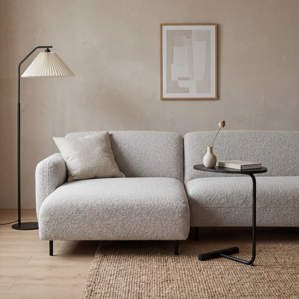 Lichte woonkamer met beige hoekbank, zwarte bijzettafel, vloerlamp en neutrale wanddecoratie