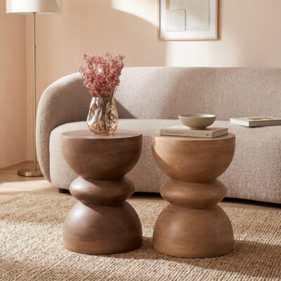 Warme woonkamer met twee sculpturale houten bijzettafels, beige bank, vaas met roze droogbloemen en vloerlamp